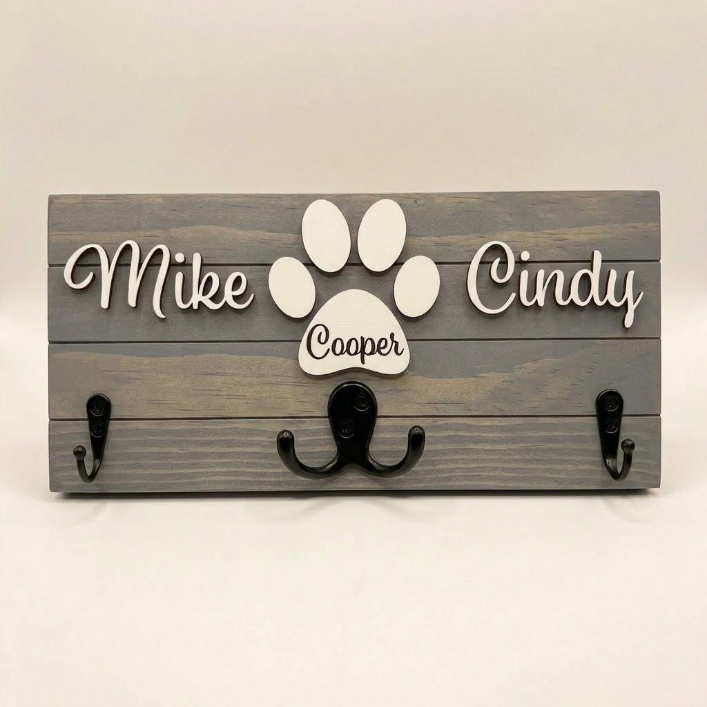 Personalized dog leash holder for wall with raised 3D names, engraved paw and black metal hooks on a stained pine board