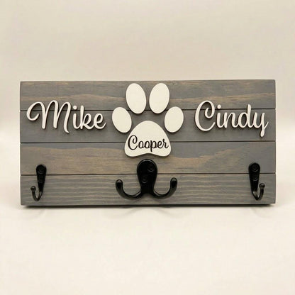 Personalized dog leash holder for wall with raised 3D names, engraved paw and black metal hooks on a stained pine board
