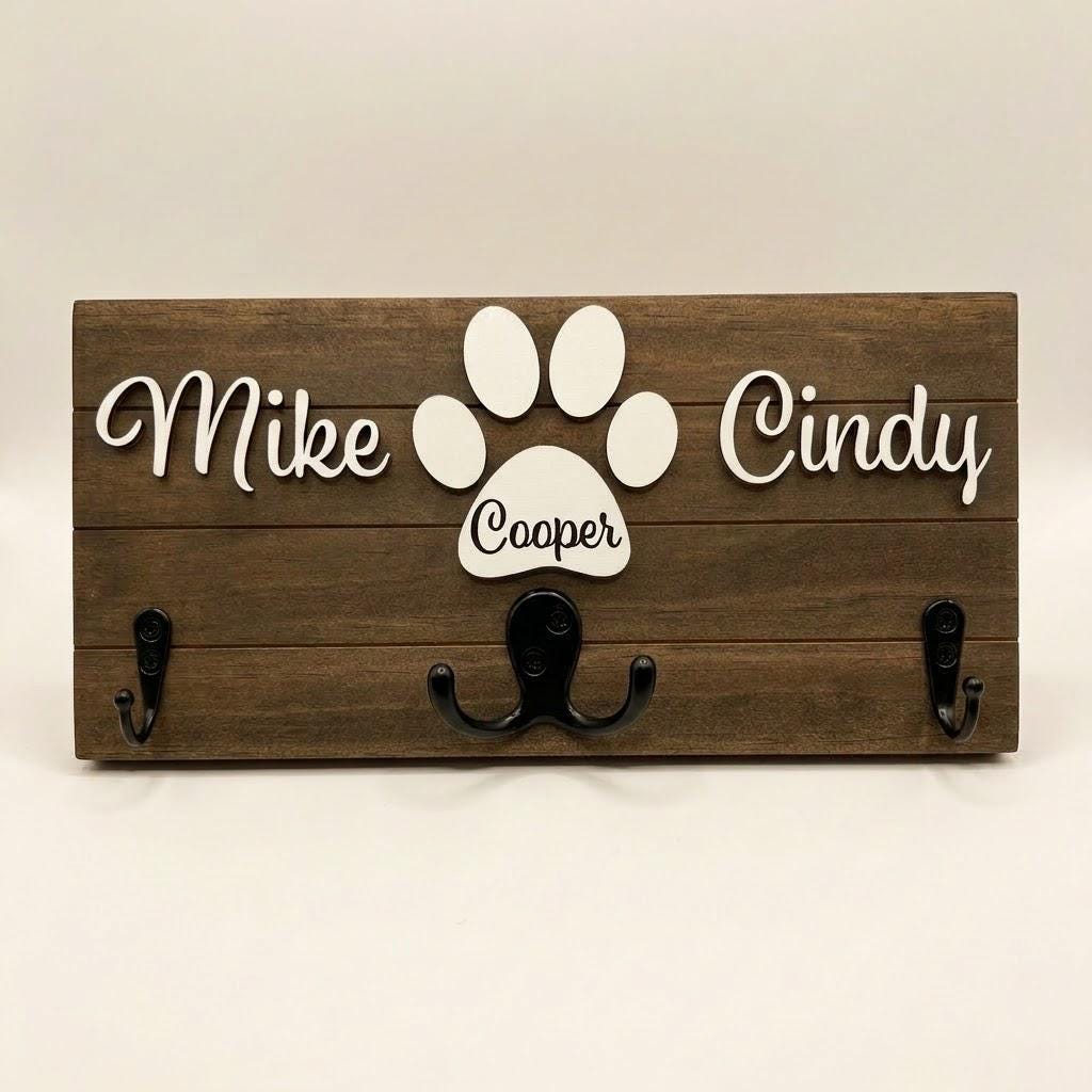 Personalized dog leash holder for wall with raised 3D names, engraved paw and black metal hooks on a stained pine board