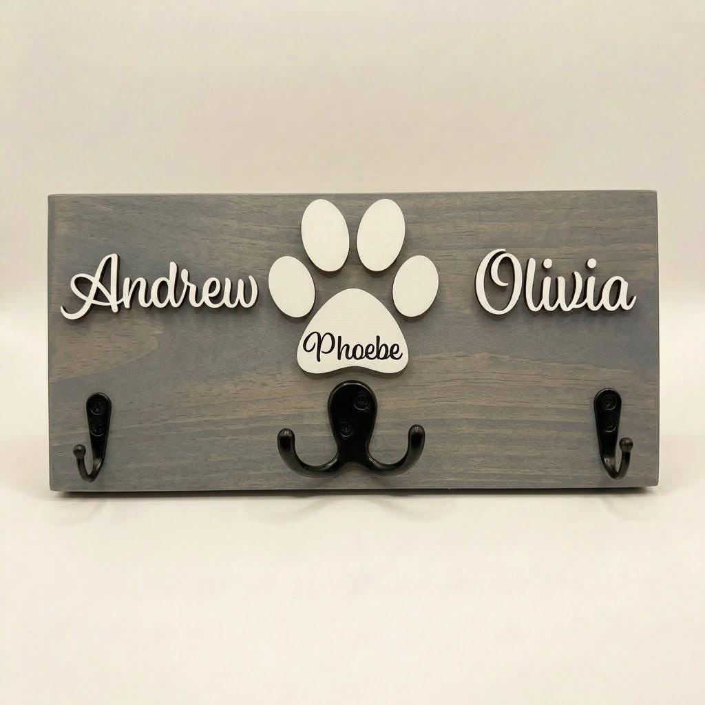 Personalized dog leash holder for wall with raised 3D names, engraved paw and black metal hooks on a stained pine board