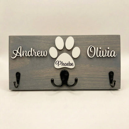 Personalized dog leash holder for wall with raised 3D names, engraved paw and black metal hooks on a stained pine board