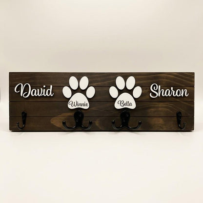 Personalized dog leash holder for wall with raised 3D names, engraved paw and black metal hooks on a stained pine board