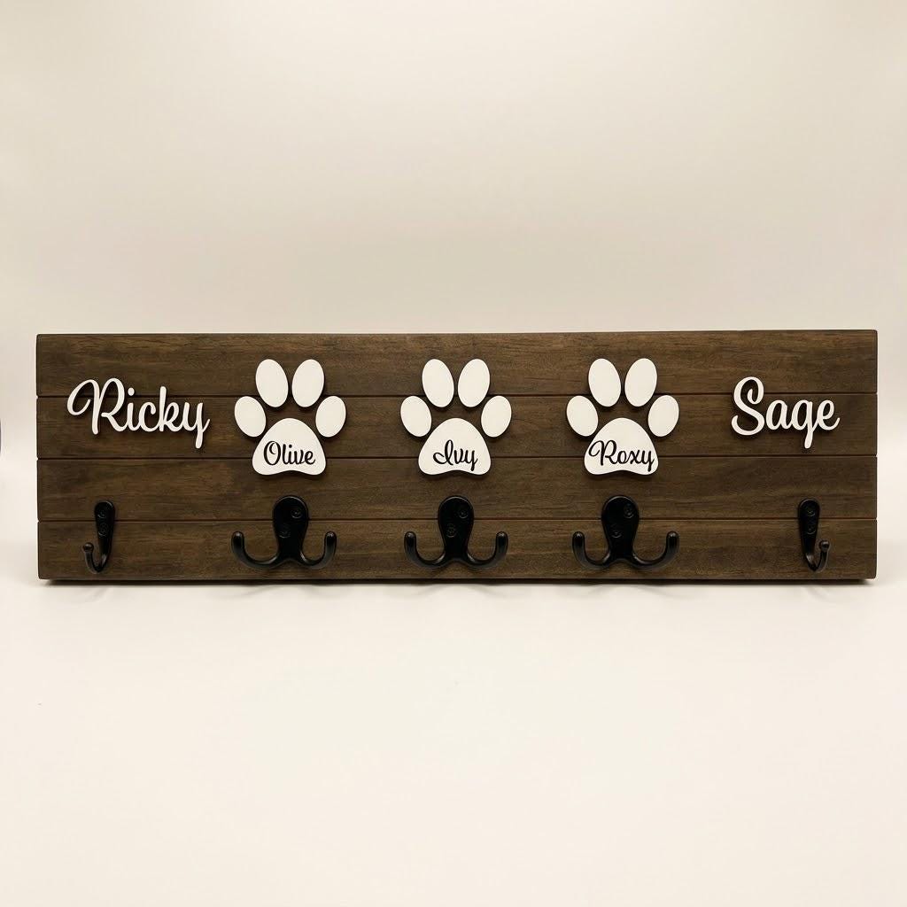 Personalized dog leash holder for wall with raised 3D names, engraved paw and black metal hooks on a stained pine board